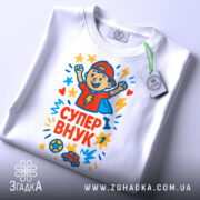 Футболка супер внук білого кольору, складена на столі, з принтом, що зображує хлопчика в червоній кепці, який радіє, оточений кольоровими зірочками, з етикеткою на зеленій стрічці, на фоні світло-сірого кольору.