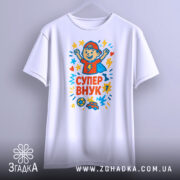 Футболка супер внук білого кольору, підвішена на вішалці, з принтом, що містить малюнок хлопчика в червоній кепці, який піднімає руки, оточений кольоровими зірочками, на фоні світло-сірого кольору.