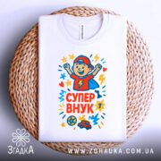 Футболка супер внук білого кольору, складена на плетеному кошику, з принтом, що зображує хлопчика в червоній кепці, який радіє, оточений зірочками та сердечками, на фоні світло-сірого кольору.