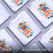 Футболка супер внук білого кольору, зображена на манекені, з принтом, що містить малюнок хлопчика в червоній кепці, який піднімає руки вгору, оточений зірочками та сердечками, на фоні світло-сірого кольору.