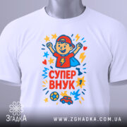 Футболка супер внук білого кольору, складена на столі, з принтом, що зображує хлопчика в червоній кепці, який радіє, оточений кольоровими зірочками, з етикеткою на зеленій стрічці, на фоні світло-сірого кольору.