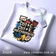 Футболка суперсили копіювання, біла, зображена зверху, складена на поверхні, фон світло-сірий, принт з текстом "Ctrl + C Ctrl + V мої суперсили" яскравих кольорів, з етикеткою на комірці.