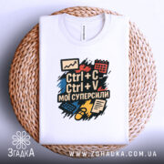 Футболка суперсили копіювання, біла, зображена зверху, складена на плетеній кошику, фон світло-сірий, принт містить текст "Ctrl + C Ctrl + V мої суперсили" на кольорових фонах з графічними елементами.