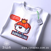 Футболка суші король, біла, складена на столі, фон світлий, принт з веселим суші персонажем у червоному капелюшку та написом 'Піддані, несіть більше соєвого соусу!', на футболці є етикетка з зеленим шнурком.