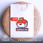 Футболка суші король, біла, складена на плетеному кошику, фон нейтральний, принт з веселим суші персонажем у червоному капелюшку та написом 'Піддані, несіть більше соєвого соусу!'.
