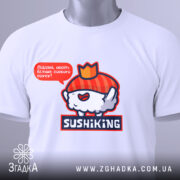 Футболка суші король, біла, складена на столі, фон світлий, принт з веселим суші персонажем у червоному капелюшку та написом 'Піддані, несіть більше соєвого соусу!', на футболці є етикетка з зеленим шнурком.
