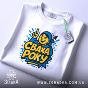 Футболка сваха року, біла, зображена зверху, на столі, фон світло-сірий, принт з жовтим текстом 'СВАХА РОКУ' та ілюстрацією усміхненої жінки, на футболці є бірка з зеленим шнурком.