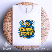 Футболка сваха року, біла, зображена зверху на плетеній кошику, фон світло-сірий, принт з жовтим текстом 'СВАХА РОКУ' та ілюстрацією усміхненої жінки, видима етикетка з написом.