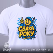 Футболка сваха року, біла, зображена зверху, на столі, фон світло-сірий, принт з жовтим текстом 'СВАХА РОКУ' та ілюстрацією усміхненої жінки, на футболці є бірка з зеленим шнурком.