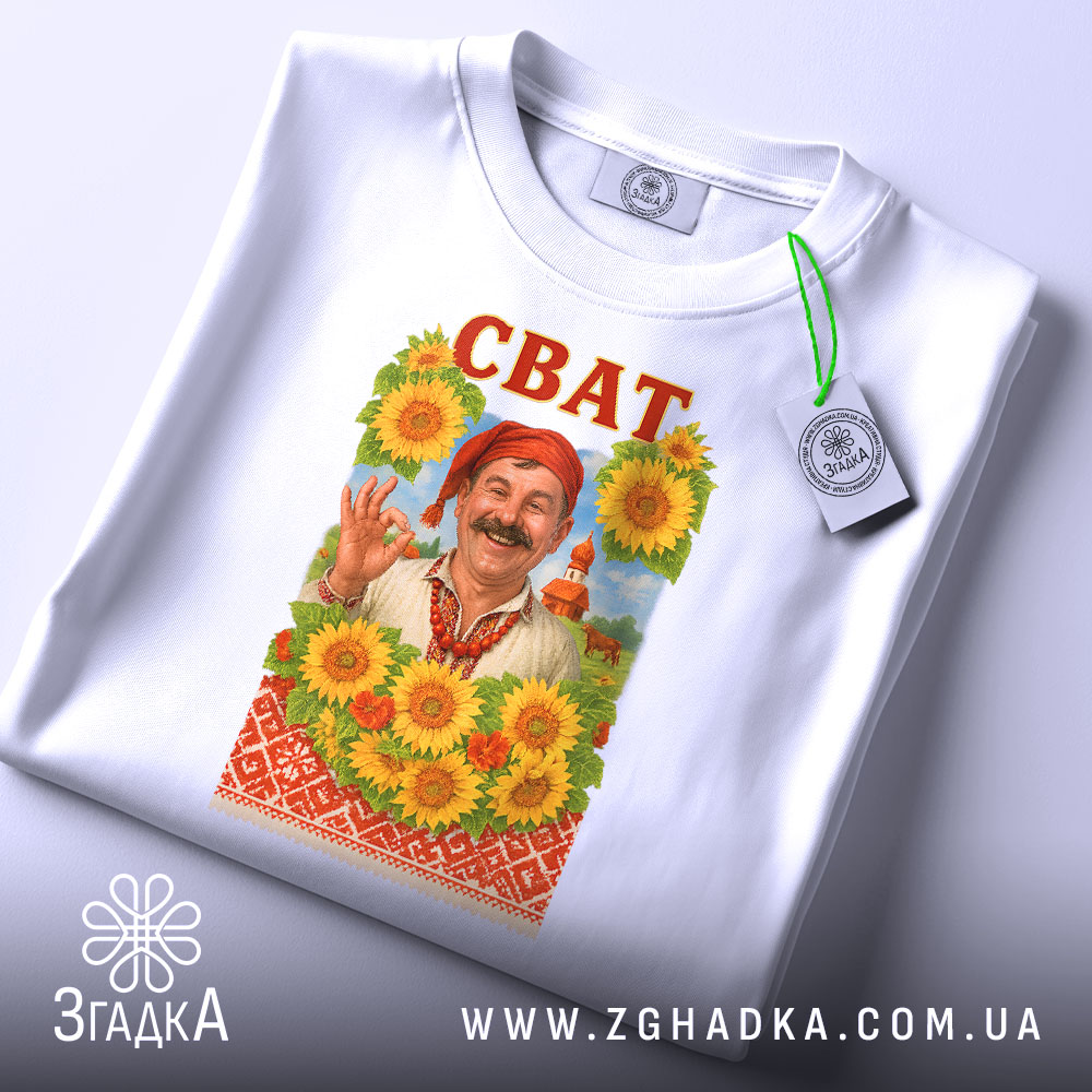 Футболка сват білого кольору, складена на поверхні, фон світло-сірий, принт з усміхненим чоловіком у традиційному вбранні на фоні соняшників, напис 'СВАТ' червоним кольором вгорі, на футболці є етикетка з зеленим шнурком.