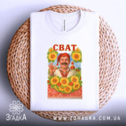 Футболка сват білого кольору, складена на плетеній кошику, фон світло-сірий, принт з усміхненим чоловіком у традиційному вбранні на фоні соняшників, напис 'СВАТ' червоним кольором вгорі.