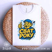 Футболка сват року, біла, складена на плетеній кошику, фон світло-сірий, принт з усміхненим чоловіком у капелюсі, який піднімає великий палець, текст "СВАТ РОКУ" яскраво-жовтого кольору.