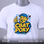 Футболка сват року, біла, складена на столі, фон світло-сірий, принт з усміхненим чоловіком у капелюсі, який піднімає великий палець, текст "СВАТ РОКУ" яскраво-жовтого кольору, на футболці є етикетка з зеленим шнурком.