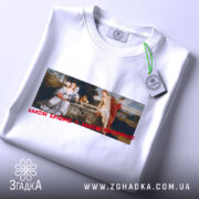 Футболка свята любов, біла, складена на поверхні, фон світло-сірий, принт з класичною картиною, текст червоним шрифтом 'AMOR SACRO Y AMOR PROFANO', на ярлику зелена стрічка.