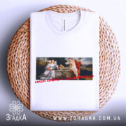 Футболка свята любов, біла, складена на кошику, фон світлий, принт з класичною картиною, текст червоним шрифтом 'AMOR SACRO Y AMOR PROFANO'.