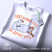 Футболка святковий гусь білого кольору, складена на плоскій поверхні, фон світло-сірий, принт з гусем у смокінгу, що лежить, з написом 'ЗА ДУПКУ КУСЬ' у яскравих кольорах, на етикетці зелена стрічка.