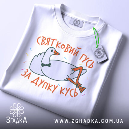 Футболка святковий гусь білого кольору, складена на плоскій поверхні, фон світло-сірий, принт з гусем у смокінгу, що лежить, з написом 'ЗА ДУПКУ КУСЬ' у яскравих кольорах, на етикетці зелена стрічка.