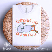 Футболка святковий гусь білого кольору, складена на плетеній кошику, фон світло-сірий, принт з гусем у смокінгу, що лежить, з написом 'ЗА ДУПКУ КУСЬ' у яскравих кольорах.