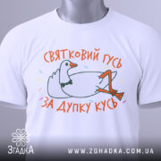 Футболка святковий гусь білого кольору, складена на плоскій поверхні, фон світло-сірий, принт з гусем у смокінгу, що лежить, з написом 'ЗА ДУПКУ КУСЬ' у яскравих кольорах, на етикетці зелена стрічка.