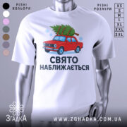 Футболка свято наближається. Біла футболка з коротким рукавом, зображення червоного автомобіля з ялинкою на даху, текст "СВЯТО НАБЛИЖАЄТЬСЯ" під малюнком, фон нейтральний, деталі принта яскраві.