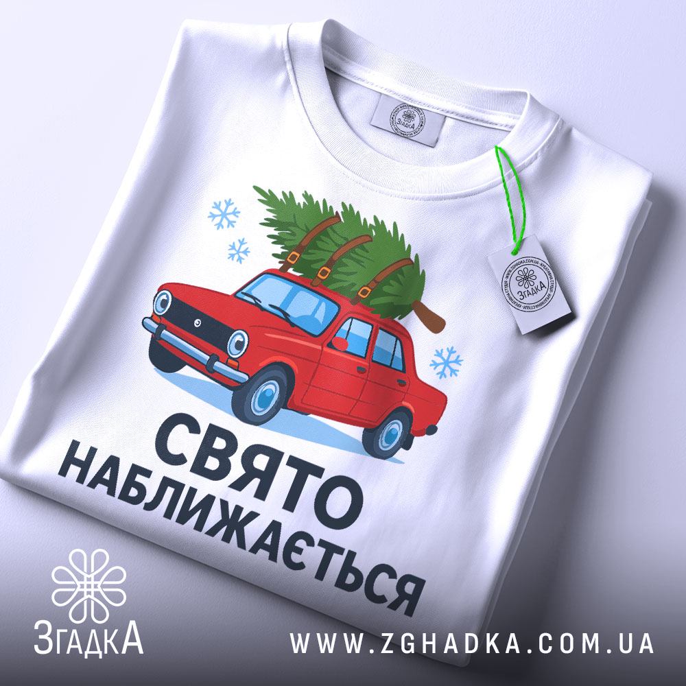 Футболка свято наближається. Біла футболка, складена на поверхні, з малюнком червоного автомобіля з ялинкою, текст "СВЯТО НАБЛИЖАЄТЬСЯ" під малюнком, на футболці є етикетка з зеленим шнурком, фон світлий.