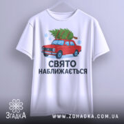 Футболка свято наближається. Біла футболка підвішена на вішалці, з малюнком червоного автомобіля з ялинкою, текст "СВЯТО НАБЛИЖАЄТЬСЯ" під малюнком, фон світло-сірий, деталі принта чіткі.