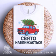 Футболка свято наближається. Біла футболка, складена на плетеній основі, з малюнком червоного автомобіля з ялинкою, текст "СВЯТО НАБЛИЖАЄТЬСЯ" під малюнком, фон світлий, деталі принта яскраві.