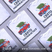 Футболка свято наближається. Біла футболка з коротким рукавом, зображення червоного автомобіля з ялинкою на даху, текст "СВЯТО НАБЛИЖАЄТЬСЯ" під малюнком, фон нейтральний, деталі принта яскраві.