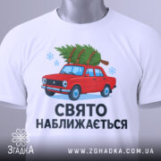 Футболка свято наближається. Біла футболка, складена на поверхні, з малюнком червоного автомобіля з ялинкою, текст "СВЯТО НАБЛИЖАЄТЬСЯ" під малюнком, на футболці є етикетка з зеленим шнурком, фон світлий.