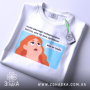 Футболка світла вже не треба. Біла футболка, складена на столі, з видимим етикеткою. Фон світлий, з легким градієнтом. Принт з ілюстрацією дівчини з рудим волоссям, текст на українській мові розташований над малюнком.