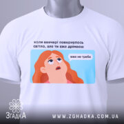 Футболка світла вже не треба. Біла футболка, складена на столі, з видимим етикеткою. Фон світлий, з легким градієнтом. Принт з ілюстрацією дівчини з рудим волоссям, текст на українській мові розташований над малюнком.