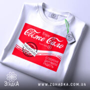 Футболка свіже сало білого кольору, складена на поверхні, з принтом червоного кольору, на якому написано "Свіже сало" та інші деталі, на футболці є зелений ярлик, фон світлий.