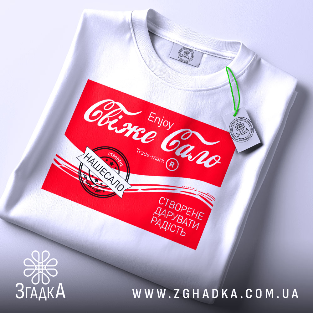 Футболка свіже сало білого кольору, складена на поверхні, з принтом червоного кольору, на якому написано "Свіже сало" та інші деталі, на футболці є зелений ярлик, фон світлий.