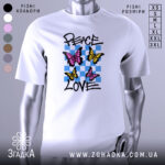 Футболка свобода любові. Білий колір, зображена на манекені з правого боку, фон світло-сірий. Принт містить три метелики: два рожевих та один жовтий, на фоні з блакитних квадратів, текст 'PEACE LOVE' написаний чорним шрифтом.