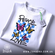 Футболка свобода любові. Білий колір, зображена на столі, ракурс зверху. Принт з трьома метеликами: два рожевих та один жовтий, на фоні з блакитних квадратів, текст 'PEACE LOVE' написаний чорним шрифтом. На футболці є етикетка з зеленим шнурком.