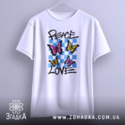 Футболка свобода любові. Білий колір, зображена на вішалці, фон світло-сірий. Принт містить три метелики: два рожевих та один жовтий, на фоні з блакитних квадратів, текст 'PEACE LOVE' написаний чорним шрифтом.