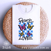 Футболка свобода любові. Білий колір, зображена на плетеному кошику, ракурс зверху. Принт містить три метелики: два рожевих та один жовтий, на фоні з блакитних квадратів, текст 'PEACE LOVE' написаний чорним шрифтом.