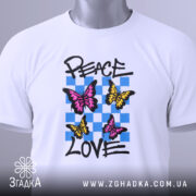 Футболка свобода любові. Білий колір, зображена на столі, ракурс зверху. Принт з трьома метеликами: два рожевих та один жовтий, на фоні з блакитних квадратів, текст 'PEACE LOVE' написаний чорним шрифтом. На футболці є етикетка з зеленим шнурком.