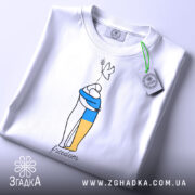Футболка свобода Україна, біла, складена на столі, з верхнього ракурсу, фон світлий, принт з двома фігурами, одна з яких тримає жовту і синю смугу, напис 'freedom' під малюнком, на комірці видна етикетка.