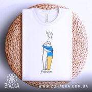 Футболка свобода Україна, біла, складена на плетеній кошику, з верхнього ракурсу, фон світлий, принт з двома фігурами, одна з яких тримає жовту і синю смугу, напис 'freedom' під малюнком.