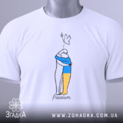 Футболка свобода Україна, біла, складена на столі, з верхнього ракурсу, фон світлий, принт з двома фігурами, одна з яких тримає жовту і синю смугу, напис 'freedom' під малюнком, на комірці видна етикетка.