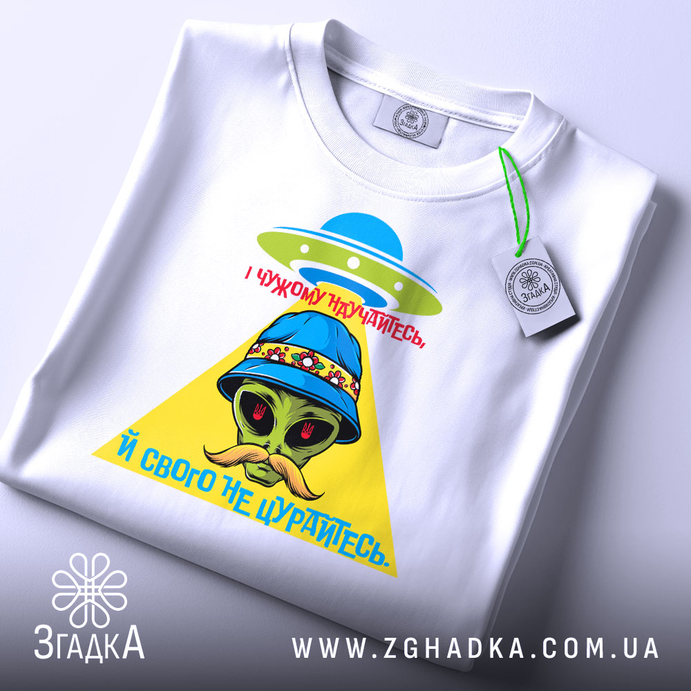 Футболка свого не цурайтесь, складена футболка білого кольору, на ній принт з прибульцем у капелюсі, жовтий трикутник та червоний текст, фон світло-сірий.