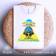 Футболка свого не цурайтесь, складена футболка білого кольору на плетеній основі, принт з прибульцем у капелюсі, жовтий трикутник та червоний текст, фон світло-сірий.