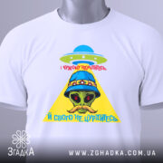 Футболка свого не цурайтесь, складена футболка білого кольору, на ній принт з прибульцем у капелюсі, жовтий трикутник та червоний текст, фон світло-сірий.