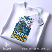 Футболка сила дронів, біла, зображена зверху, фон світло-сірий, принт з військовим, що керує дронами, з написом 'СИЛА ДРОНІВ' у жовтому кольорі, з етикеткою на шнурку.