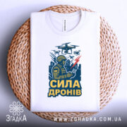Футболка сила дронів, біла, зображена зверху на плетеній кошику, фон світло-сірий, принт з військовим, що керує дронами, з написом 'СИЛА ДРОНІВ' у жовтому кольорі.
