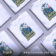 Футболка сила дронів, біла, зображена на манекені з блискучою поверхнею, фон світло-сірий, принт з військовим, що керує дронами, з написом 'СИЛА ДРОНІВ' у жовтому кольорі.