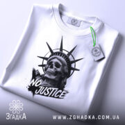 Футболка сила меседжу: біла футболка, складена на поверхні, принт з черепом та короною свободи, ракурс зверху, фон світло-сірий, на футболці є етикетка з зеленим шнурком.