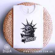 Футболка сила меседжу: біла футболка, складена на плетеній кошику, принт з черепом та короною свободи, ракурс зверху, фон світло-сірий, деталі принта чорного кольору з написом 'NO JUSTICE'.
