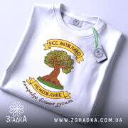 Футболка сила можливостей. Біла футболка, складена на плоскій поверхні, з видимим етикеткою. Принт з деревом та написом розташований по центру, фон світло-сірий, а футболка має округлий виріз.